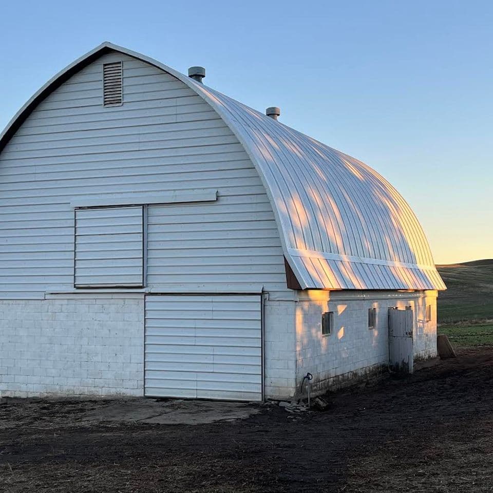 White barn with curved roof under a clear sky at sunset, surrounded by farmland.