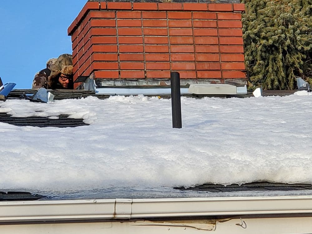 Person inspecting snow-covered roof near chimney on a sunny day.
