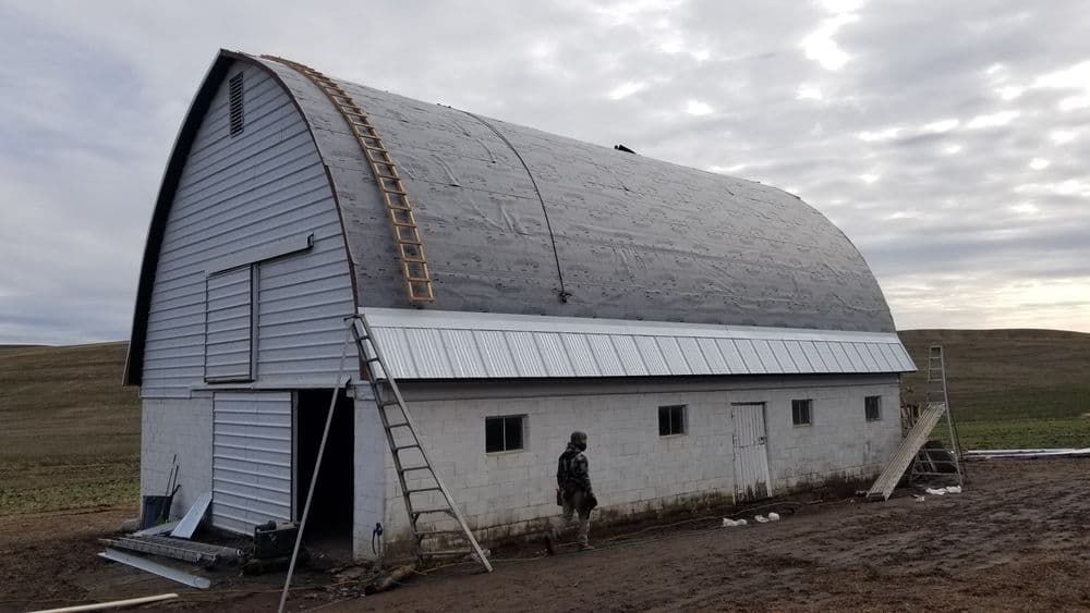 Man working on a renovated barn with a curved roof and ladders against a rural backdrop.