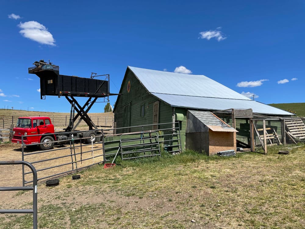 Green barn with silver roof, red truck, and equipment in rural landscape under blue sky.