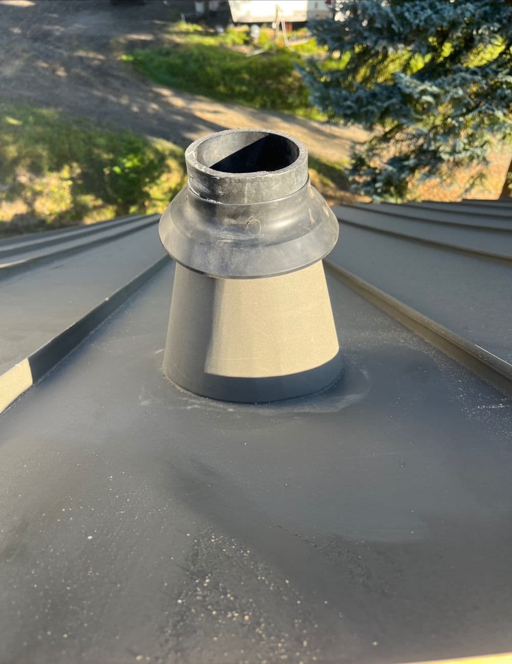 Roof ventilation pipe on a sloped metal roof, surrounded by tree foliage and gravel ground.