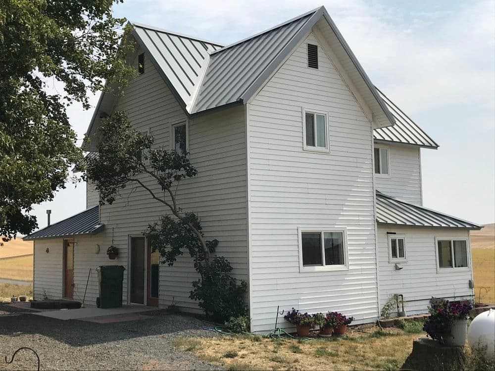 White farmhouse with metal roof, surrounded by greenery and flowers, in a rural setting.