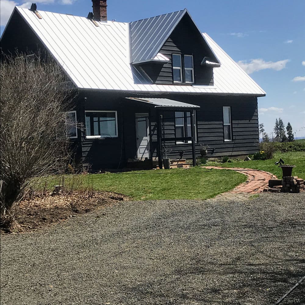 Black farmhouse with a metal roof, green lawn, and gravel driveway on a sunny day.